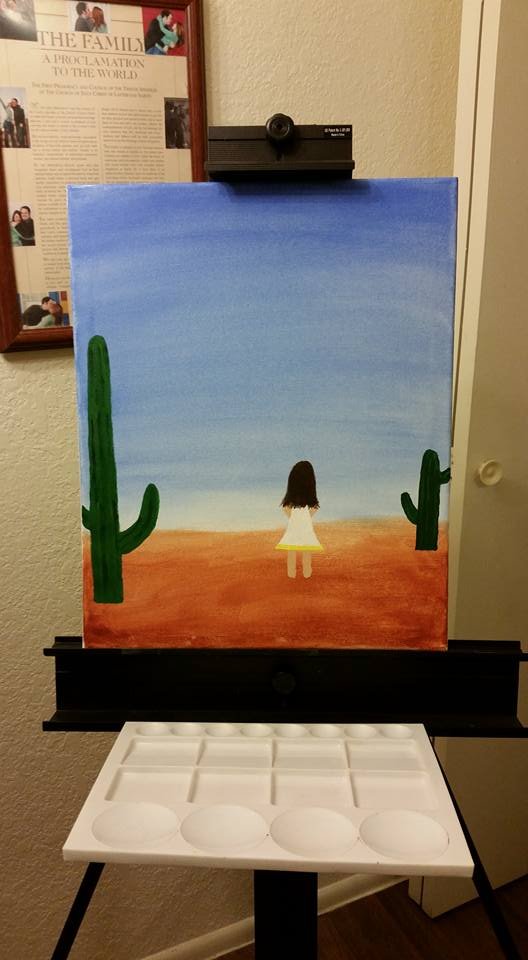 a painting on an easel of a girl wearing a white dress with a yellow ribbon at the bottom staring ahead so you only see her long brown hair. She is standing in a desert with saguaro cacti